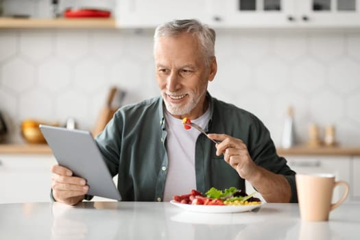 Smiling Senior Man Using Digital Tablet While Having Lunch In Kitchen