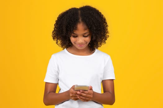 Positive adolescent black girl in white t-shirt, uses phone to chat in social networks