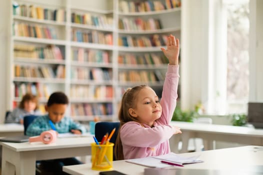 Smart female elementary school student raising her hand, ready to answer the teacher's questions in classroom interior