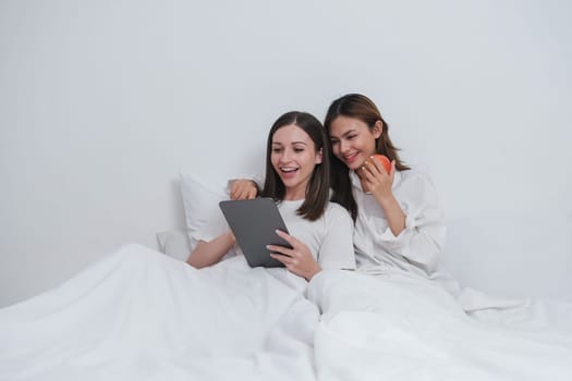 LBGT Couple of cute lesbian women waking up in the morning on white bed in bedroom while laughing and looking at tablet together