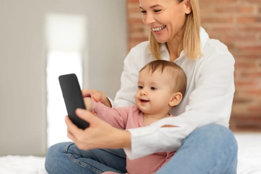 Happy european mother and infant baby girl using mobile phone playing educational games online or watching cartoons
