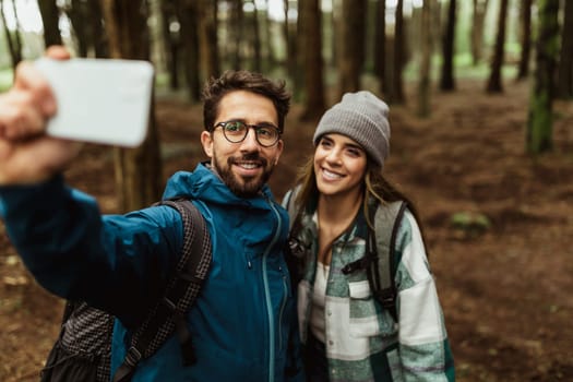 Cheerful young caucasian couple in jackets in forest, make selfie on smartphone, enjoy vacation