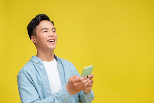Excited young Asian man  using smartphone and doing winner gesture isolated on yellow background