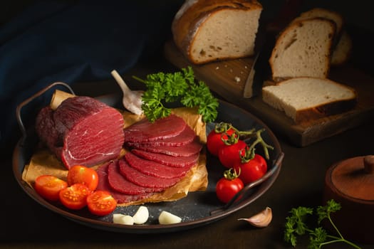 Whole and sliced bresaola on a metal round tray with tomatoes, garlic and bread.