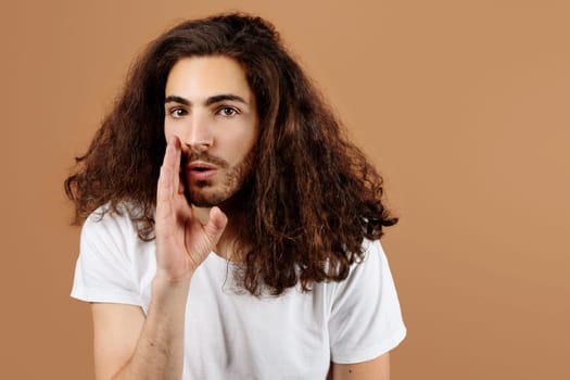 Young guy whispering secret holding hand near mouth, beige background