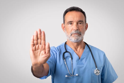 Serious confident caucasian senior doctor surgeon in blue uniform making stop sign with hand