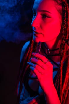 Caucasian girl with dreadlocks smokes a vape in red blue light.