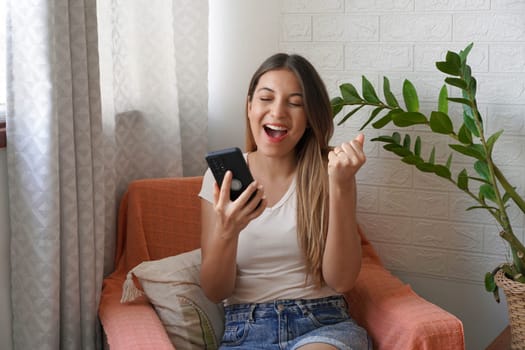 Excited happy young Brazilian woman holding cellphone sitting on armchair at home. Happy satisfied female raises her fist up with mobile phone in her hand. 