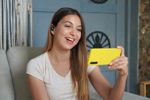 Brazilian woman watching broadcasting on video sharing platform sitting on sofa. Girl video calls with her smartphone at home. 