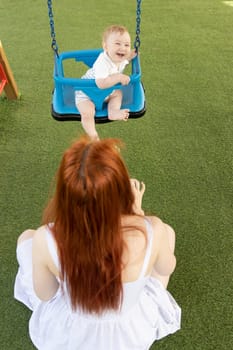 A little funny boy on swings on an outside playground with his ginger mother