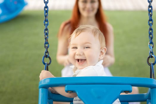 A little laughing baby on swings on an outside playground with his ginger mother