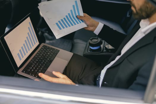 Close up of male analyst sitting on car backseat and working with documents use laptop