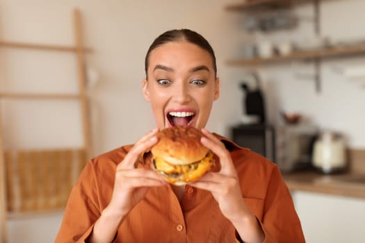Joyful woman holding a juicy burger in kitchen