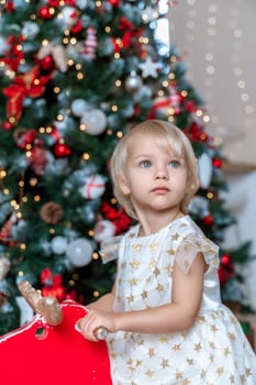 little girl Christmas tree. Blonde 2 years old in a dress sits on a wooden red horse in a room decorated for christmas and new year. Photo under the tree.