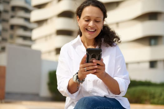 Beautiful smiling woman expressing positive emotions while using smartphone outdoors, sitting against cityscape background