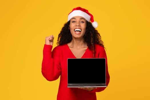 Overjoyed black woman in Santa hat showing laptop with blank screen and gesturing yes sign