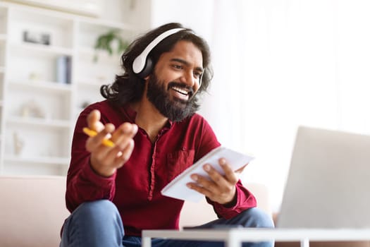 Indian guy attending webinar or online course, bearded and long-haired millennial man sitting on couch, using wireless headset and laptop, taking notes in notepad, home interior. E-education