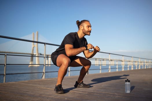 young caucasian man enjoys outdoor fitness workout squats at pier