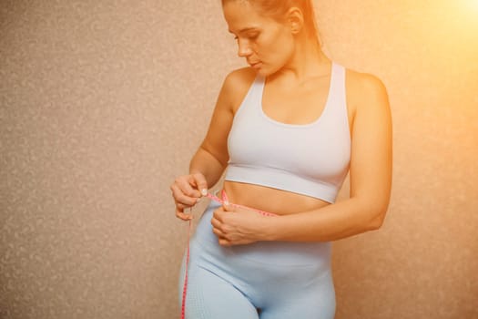 Cropped view of slim woman measuring waist with tape measure at home, close up. European woman checking the result of diet for weight loss or liposuction indoors