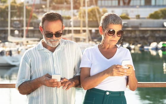 Senior husband and wife immersed in smartphones at pier outdoors