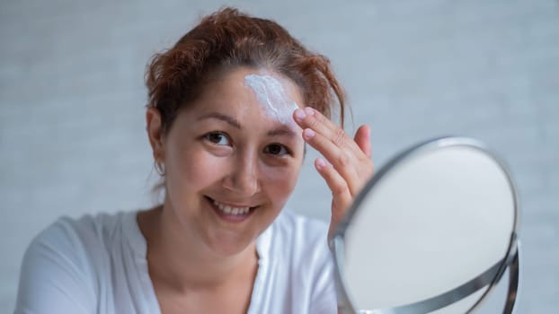 Portrait of a Caucasian woman with vitiligo uses sunscreen. A girl with a white pigment spot on her forehead looks in the mirror and is smeared with cream.