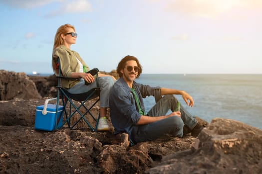 Portrait Of Man And Woman Resting In Camping Chairs On Beach Rocks