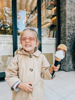 Smiling little girl with a big ice cream cone in her hand stands near the building on the street. High quality photo