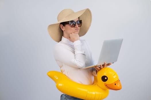 Caucasian woman works at a laptop in sunglasses, a hat and a duck in a swimming ring. Office worker dreaming of vacation.