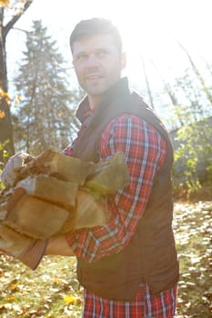 A handsome lumberjack holding a pile of wood he has collected for a cosy fire to warm a cabin in the woods. Attractive macho man carrying wood to burn while living off the grid with no worries