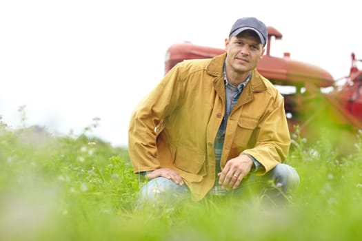 Being a farmer is part of who I am. Portrait of a farmer kneeling in a field with his tractor parked behind him.