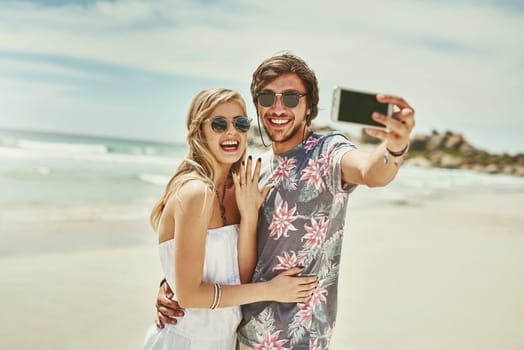 Guess whos engaged. an affectionate young couple taking selfies after their engagement on the beach.