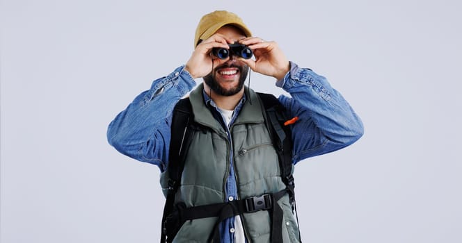 Hiking, backpack and happy man with binocular search in studio for travel, freedom or adventure on grey background. Camping, journey and backpacker with explore equipment for bird watching in nature