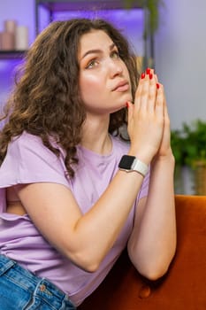 Young woman girl praying sincerely with folded arms asking God for help begging apology on home sofa