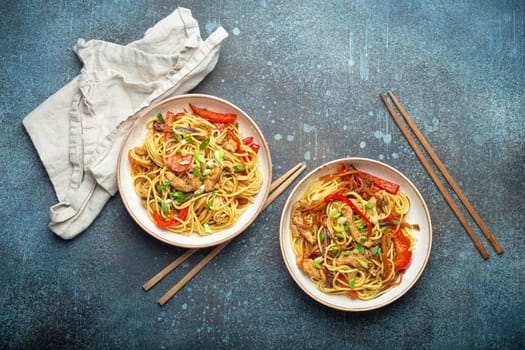 Two bowls with Chow Mein or Lo Mein, traditional Chinese stir fry noodles with meat and vegetables, served with chopsticks top view on rustic blue concrete background
