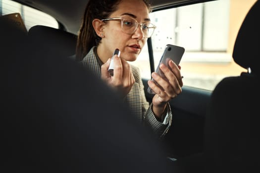 Phone, lipstick and a business woman in a taxi to travel in the city for work while multitasking. Face, mobile and makeup with a young female employee in a car or cab as a passenger for transport