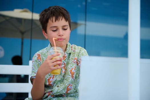 Hispanic handsome elementary age student boy in summer shirt, drinks fresh orange juice and studies online using laptop