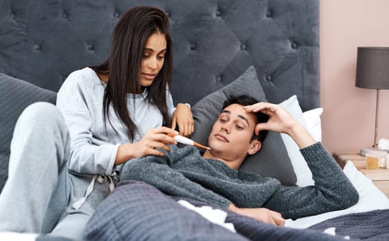 When quarantine turns your spouse into your primary care physician. a young woman taking her husband’s temperature while recovering from an illness in bed at home.
