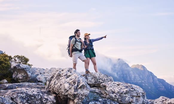 Mountain, trekking and view, man with woman on peak for adventure in nature, rock climbing and travel. Outdoor hiking, couple on cliff and relax in scenic clouds for natural journey, pointing at sky.