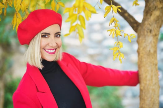 Excited young trendy woman standing near autumn tree and smiling at camera