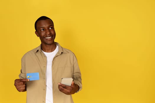 Smiling pensive millennial black guy use phone and credit card, dream, think
