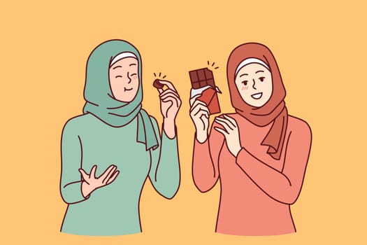 Two arab women eat chocolate and rejoice in rush of hormone of happiness, dressed in islamic clothes