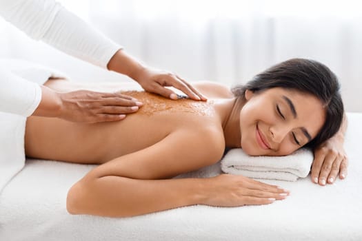 Relaxed young indian lady having skin scrubbing procedure at spa salon, closeup. Spa therapist applying exfoliating body mask on sleeping eastern woman back at luxury spa, body care concept