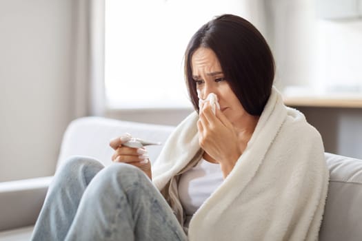 Sick Young Woman Blowing Runny Nose And Looking At Thermometer At Home