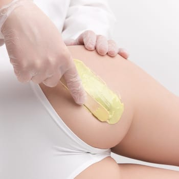 Closeup waxing procedure, depilation with hot wax in professional beauty salon. Hands in gloves applying green hot wax on buttocks of woman using spatula. Studio shot. Part of photo series.