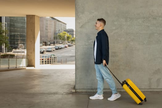 Cheerful millennial caucasian man in casual with suitcase go at plane or train at city station