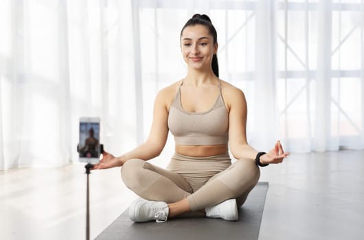 Positive woman yoga instructor meditating and recording video on smartphone