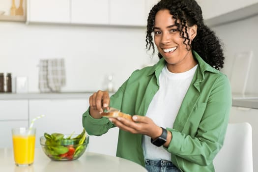 Black lady smiles making sandwich for breakfast sitting at kitchen