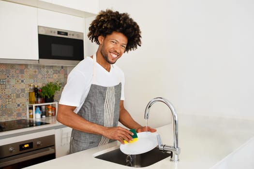 Happy black man washing dishes at sink in modern kitchen