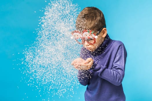 Child boy blowing snow out of his hands on blue background. Christmas and New Year holidays concept. Generation alpha and gen alpha children. Copy space