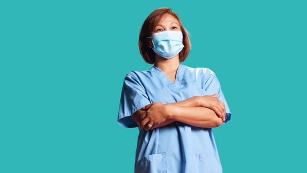 Lively healthcare specialist wearing protective medical face mask to stop virus spreading. Close up of happy smiling nurse in professional uniform with arms folded, isolated over studio background